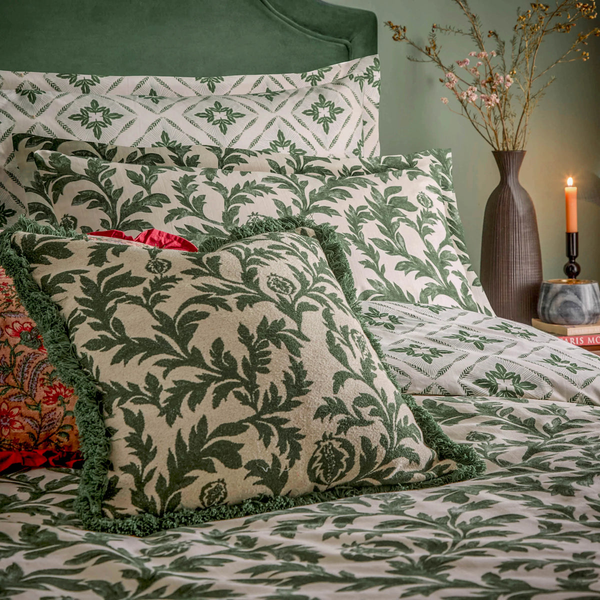 Close view of stacked pillows showing the diamond geometric reversible pattern, the ivy print Oxford border pillowcases and a green fringed ivy print cushion, with a red paisley cushion peeking out and a lit candle beside a vase on a bedside table – Kumperi