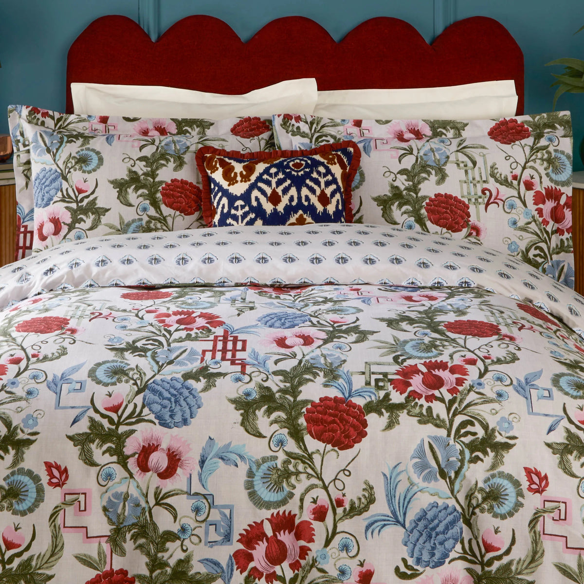 Close view of the bed showing the red and blue floral print duvet cover and reversible paisley-drop pillowcases, with a navy Ikat print cushion, set against a scalloped red velvet headboard and teal wall – Kumperi
