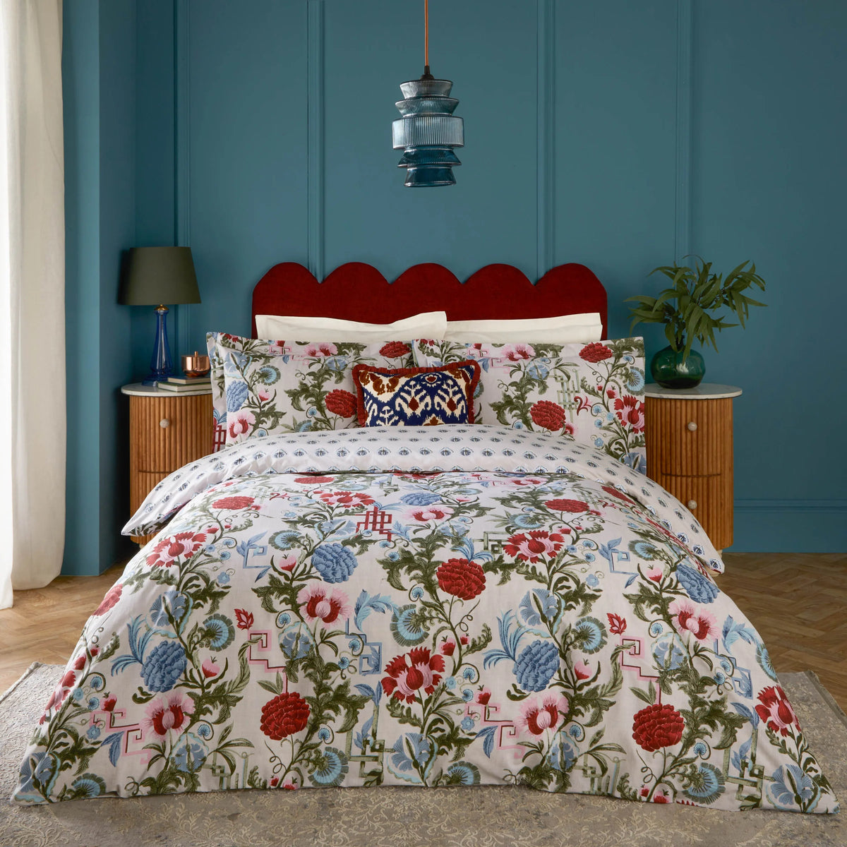 Bed dressed in the redcurrent and French blue floral duvet cover set with matching pillowcases and a navy paisley Ikat print cushion, set against a scalloped red velvet headboard, teal panelled wall, a hanging pendant light and a potted plant – Kumperi