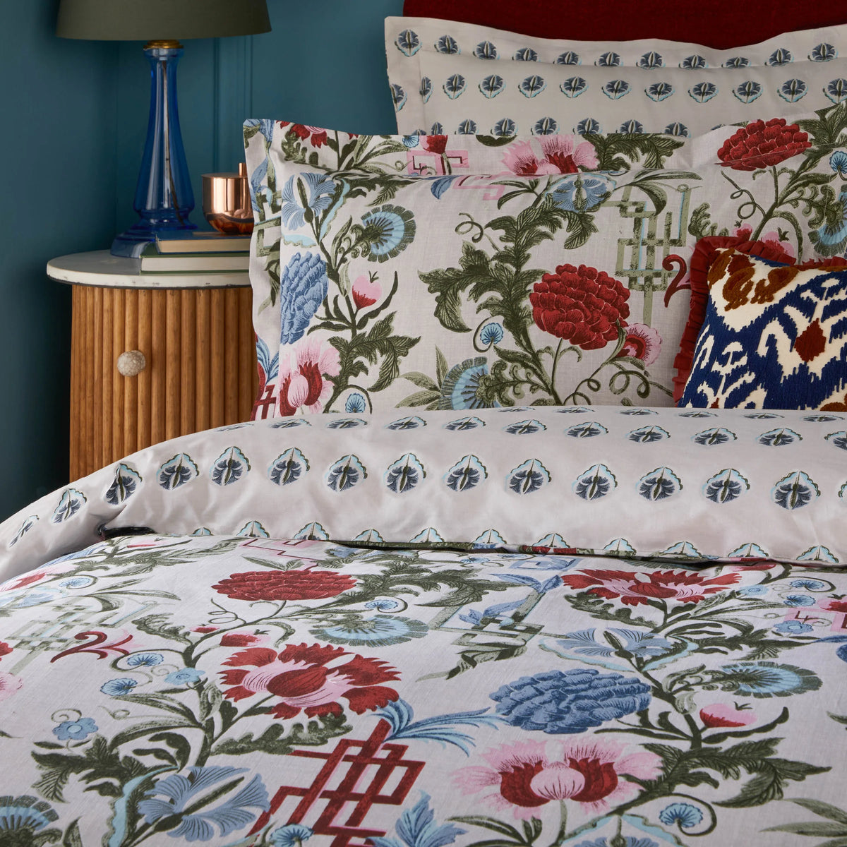 Close-up of the floral print pillowcase showing red carnation blooms, blue thistles and green foliage, layered with the reversible paisley-drop print pillowcase and a navy Ikat cushion, styled beside a bamboo bedside table with a blue glass lamp – Kumperi