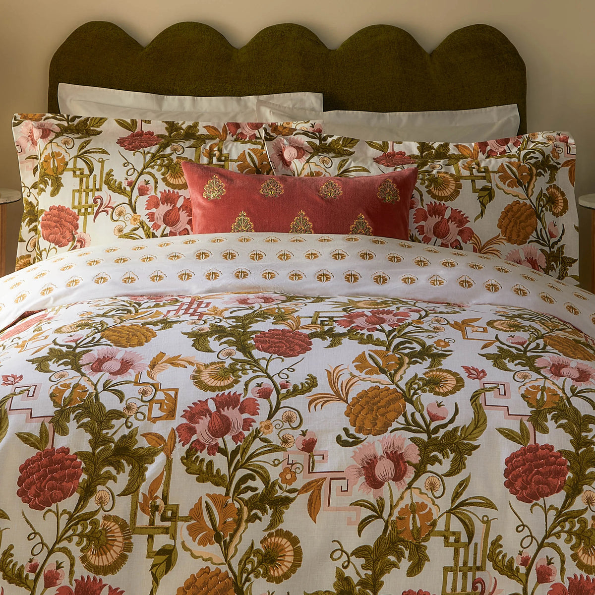 Close-up detail of the russet duvet cover and pillowcases showing large red and pink carnation-style blooms, dusky pink thistles and olive green foliage with fretwork motifs, alongside a plain rust velvet cushion – Kumperi