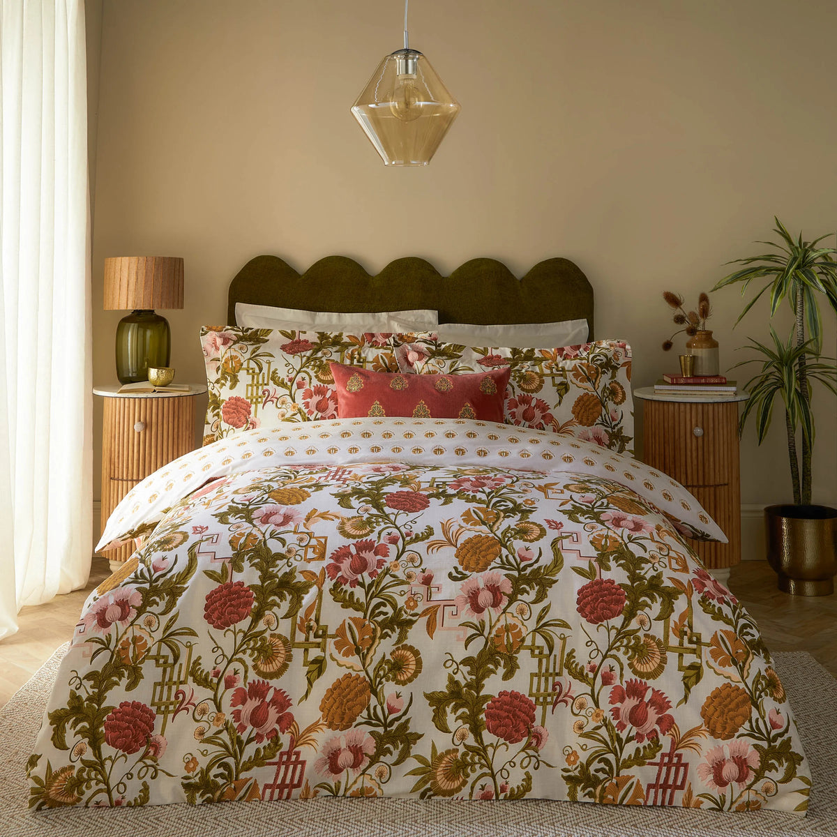 Bed dressed in the russet floral duvet cover set with matching Oxford border pillowcases and a plain rust velvet cushion, styled against a scalloped olive green velvet headboard with a hanging amber glass pendant light, rattan bedside tables, a potted plant and dried flower arrangement – Kumperi