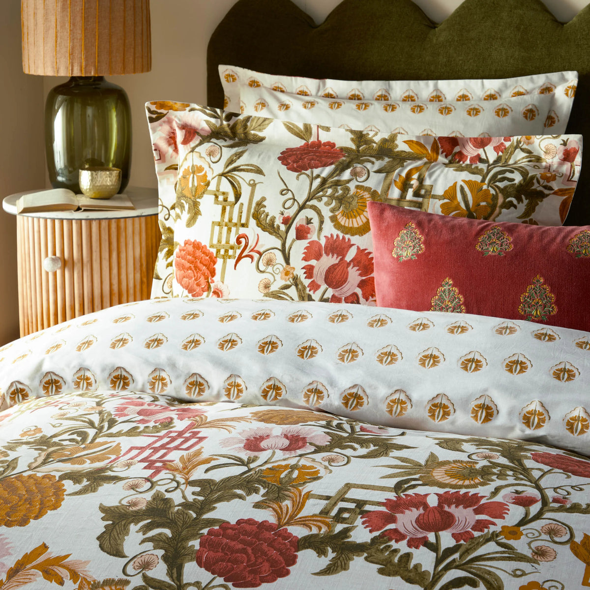 Close-up of the russet floral print Oxford border pillowcases and a plain rust velvet cushion with block-print motifs, propped against an olive green scalloped headboard beside a glass table lamp and open book on a rattan bedside table – Kumperi