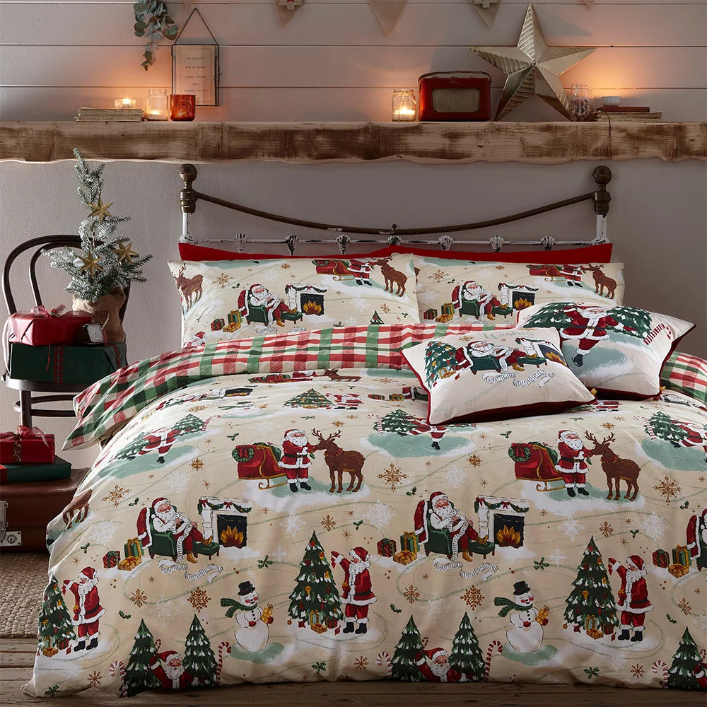 Bed dressed in the Jolly Santa Christmas duvet cover set with matching pillowcases, a buffalo check reversible pillowcase, and a small Santa print cushion, styled against a white panelled wall with a wooden mantel shelf, candles, a gold star ornament and a small decorated Christmas tree with gifts – Kumperi