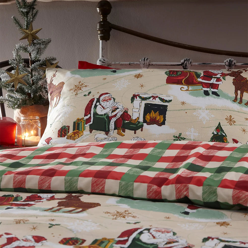 Close-up of the printed pillowcase showing Santa relaxing by the fireplace with his gift list, a reindeer and sleigh, layered against the red, green and cream buffalo check reverse, styled beside a small decorated Christmas tree and a lit candle – Kumperi
