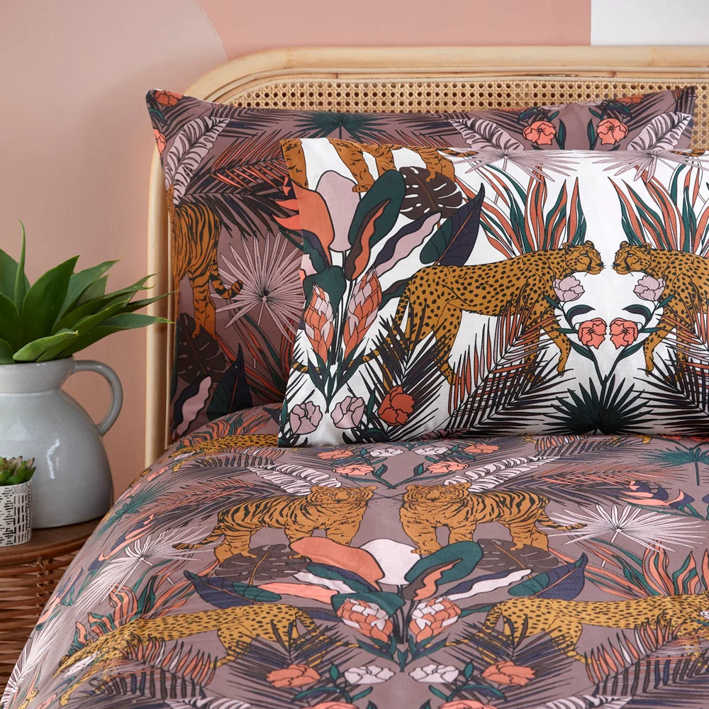 Close-up detail of the reversible pillowcases showing mirrored cheetahs and tigers among monstera leaves and coral florals, one in cocoaberry tones and one in white, propped against a rattan headboard beside a potted plant – Kumperi