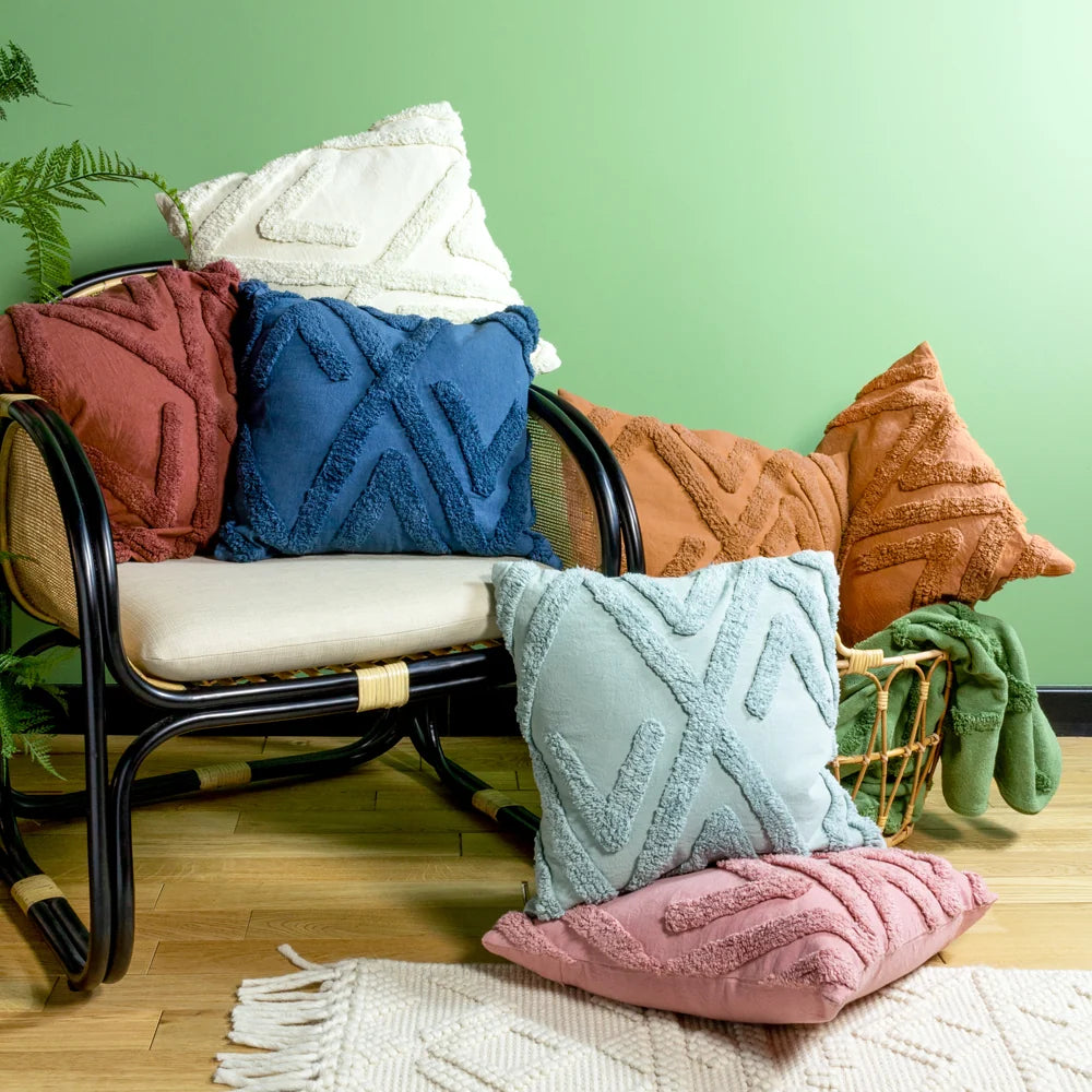 Red tufted geometric cushion styled on a rattan bench alongside matching navy, cream, terracotta, sage green and pink tufted cushions, with a folded green throw in a woven basket against a green wall – Kumperi