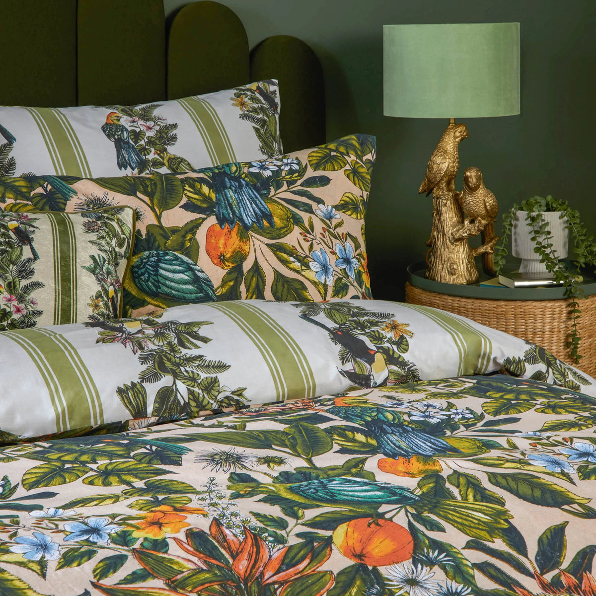 Close-up detail of the peach pillowcase print showing a blue and teal parrot among monstera leaves, citrus fruits and blue florals, layered beside the green striped toucan duvet cover fabric – Kumperi