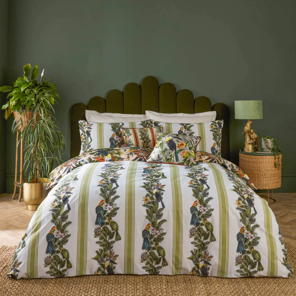 Angled view of the bed showing the olive green striped toucan duvet cover with peach botanical pillowcases and a striped cushion, set against an olive green scalloped headboard with a rattan side table, gold parrot lamp and potted plant nearby – Kumperi