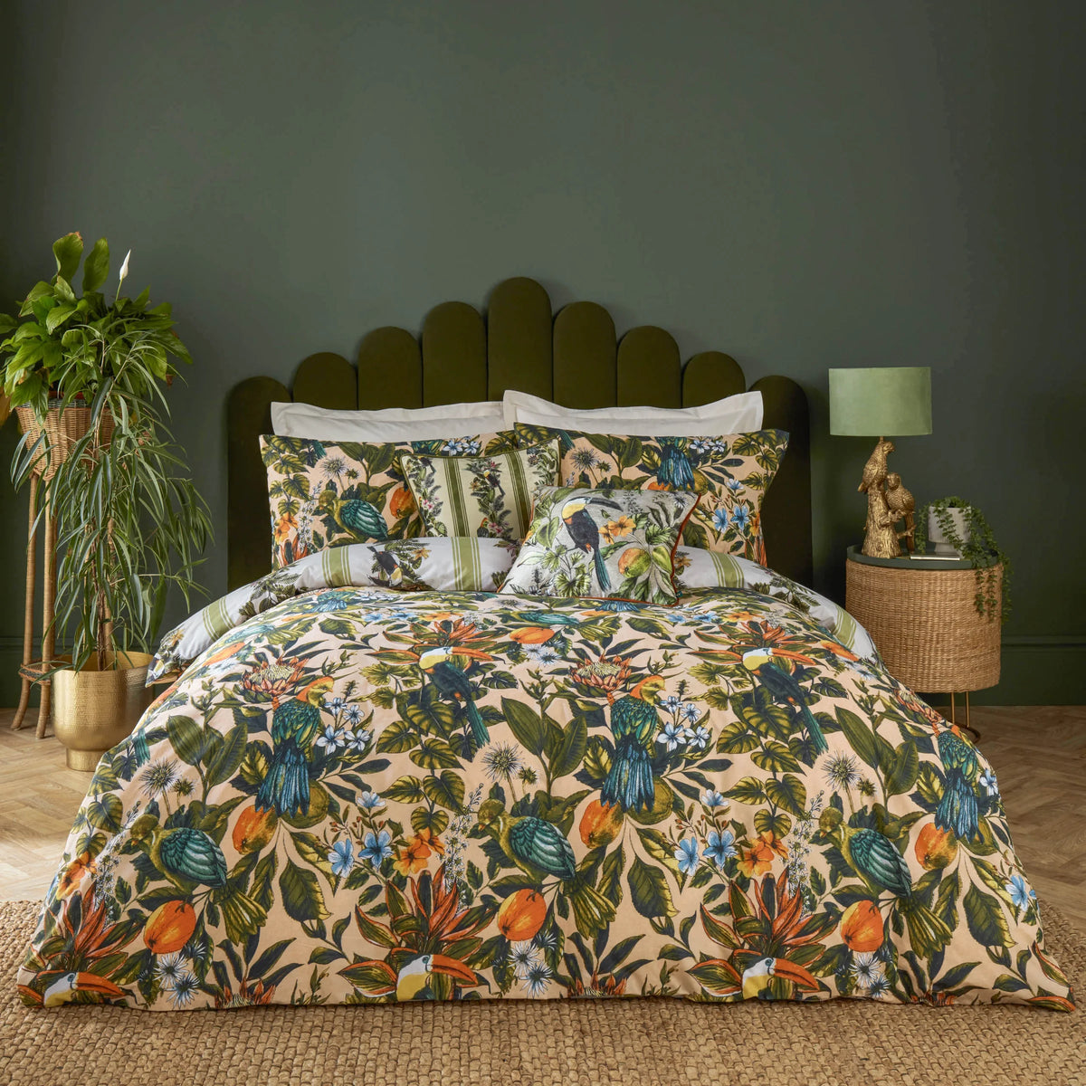 Bed dressed in the multi tropical toucan and parrot print duvet cover with peach pillowcases showing the same birds and botanical print, set against an olive green scalloped velvet headboard, styled with a rattan side table, a gold parrot lamp and a large potted plant – Kumperi