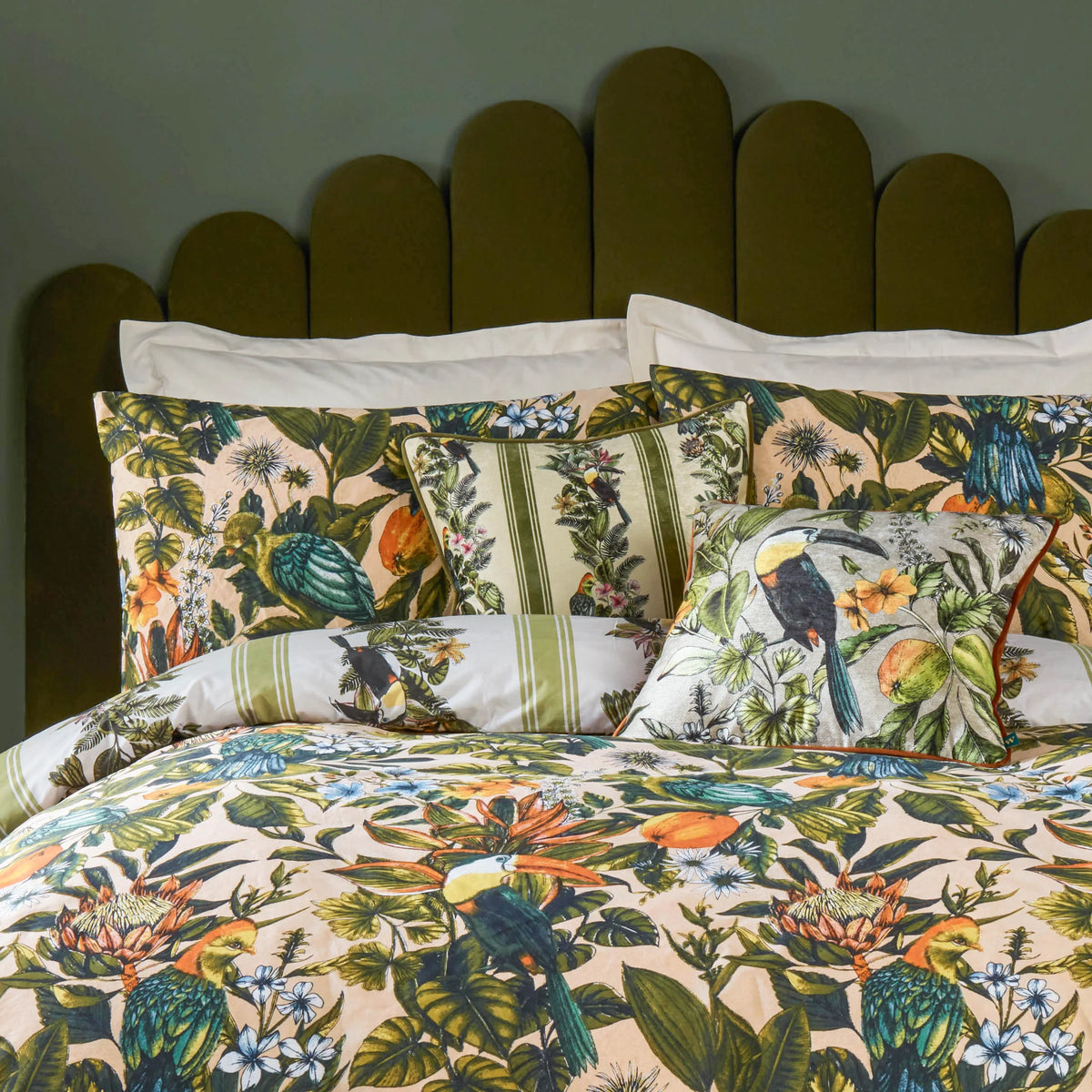 Close view of layered peach botanical print pillowcases and a green striped cushion with a toucan motif, propped against an olive green scalloped headboard, with the striped duvet cover in the foreground – Kumperi