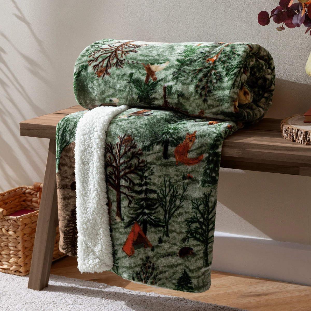 Oak green woodland print throw rolled and draped over a wooden bench, showing a fox, deer and pine trees alongside the cream sherpa fleece reverse, styled beside a woven basket and a vase of dried eucalyptus – Kumperi