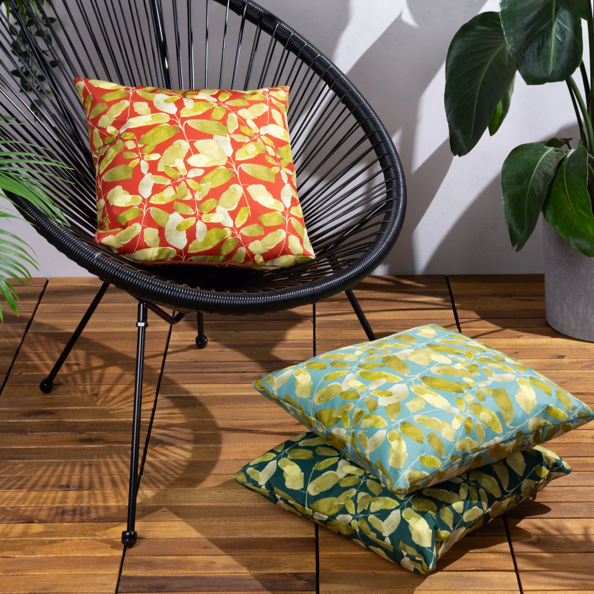 Brick red leaf print cushion styled on a black woven outdoor chair, with matching teal and dark green leaf print cushions stacked on wooden decking nearby, surrounded by potted plants – Kumperi