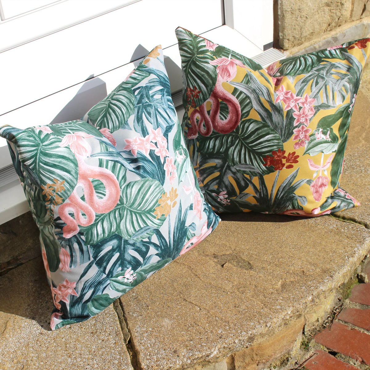 Mustard tropical snake print cushion propped on a stone doorstep beside a matching cushion in a pale grey-green tropical colourway, both featuring the pink snake motif among green leaves – Kumperi