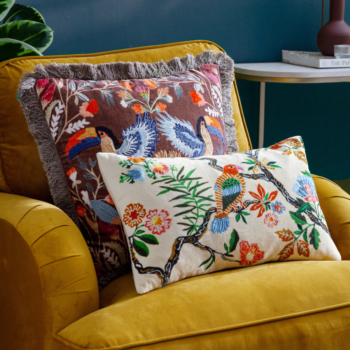 Mirrored toucan embroidered cushion with grey fringe trim styled on a mustard yellow velvet armchair alongside a smaller cream embroidered bird cushion, set against a dark teal wall with a potted plant and side table nearby – Kumperi