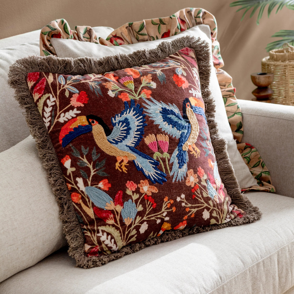Mirrored toucan embroidered cushion with grey fringe trim propped on a cream sofa beside a folded floral fringed throw, styled in a warm neutral living room setting – Kumperi