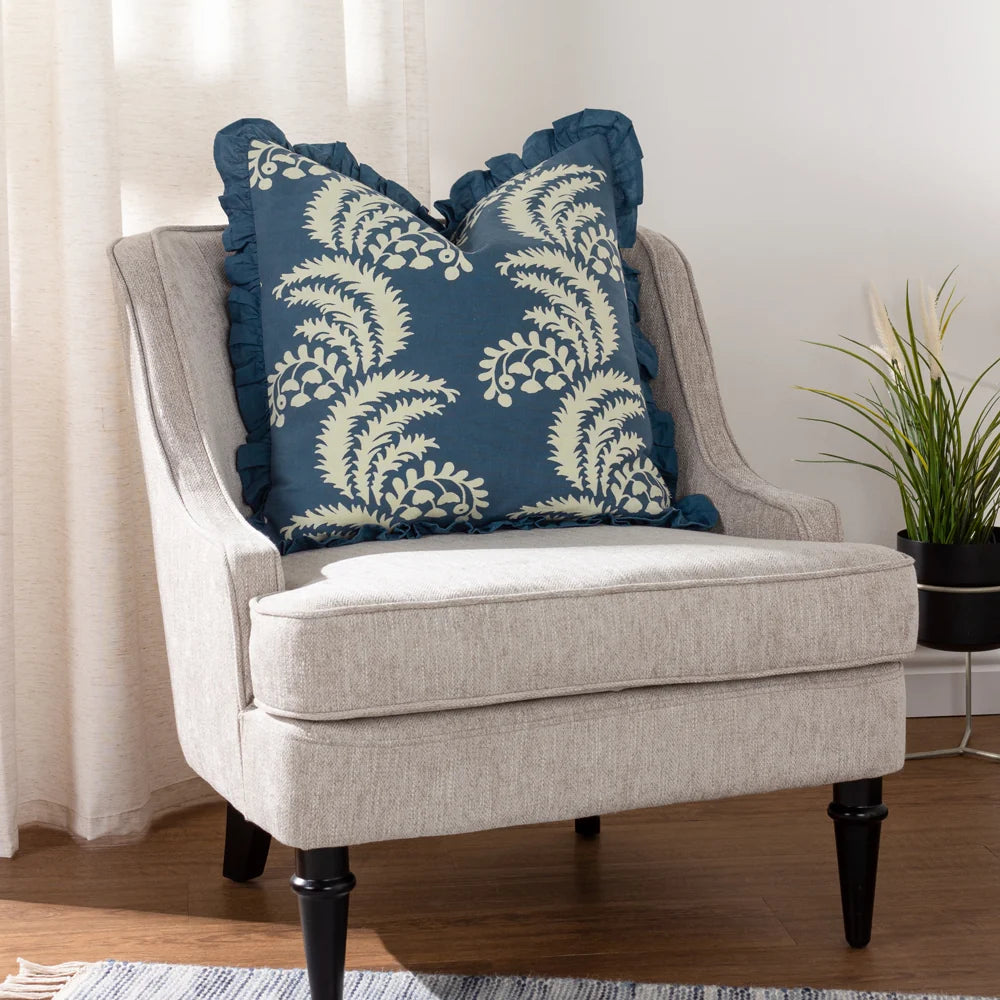 French blue fringed floral cushion styled on a beige upholstered armchair with black tapered legs, set against a cream curtain and a potted plant in the background – Kumperi