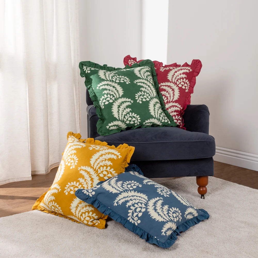 French blue fringed floral cushion shown on a rug alongside matching green, red and mustard yellow fringed cushions in the same fern and floral print, styled on and around a navy velvet armchair – Kumperi