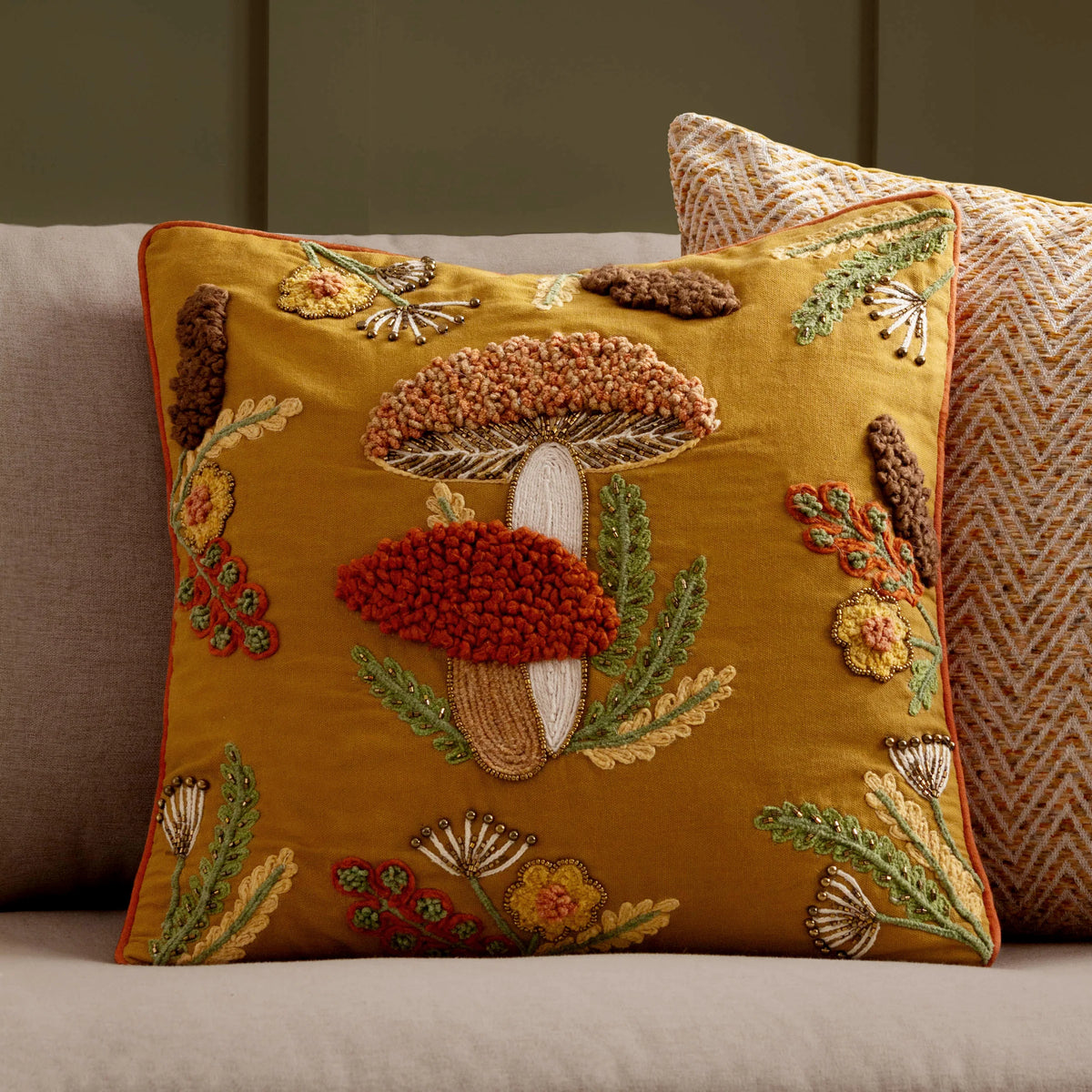 Mustard gold mushroom embroidered cushion propped against a grey sofa corner beside a cream and orange herringbone cushion, set against an olive green panelled wall with framed art above – Kumperi