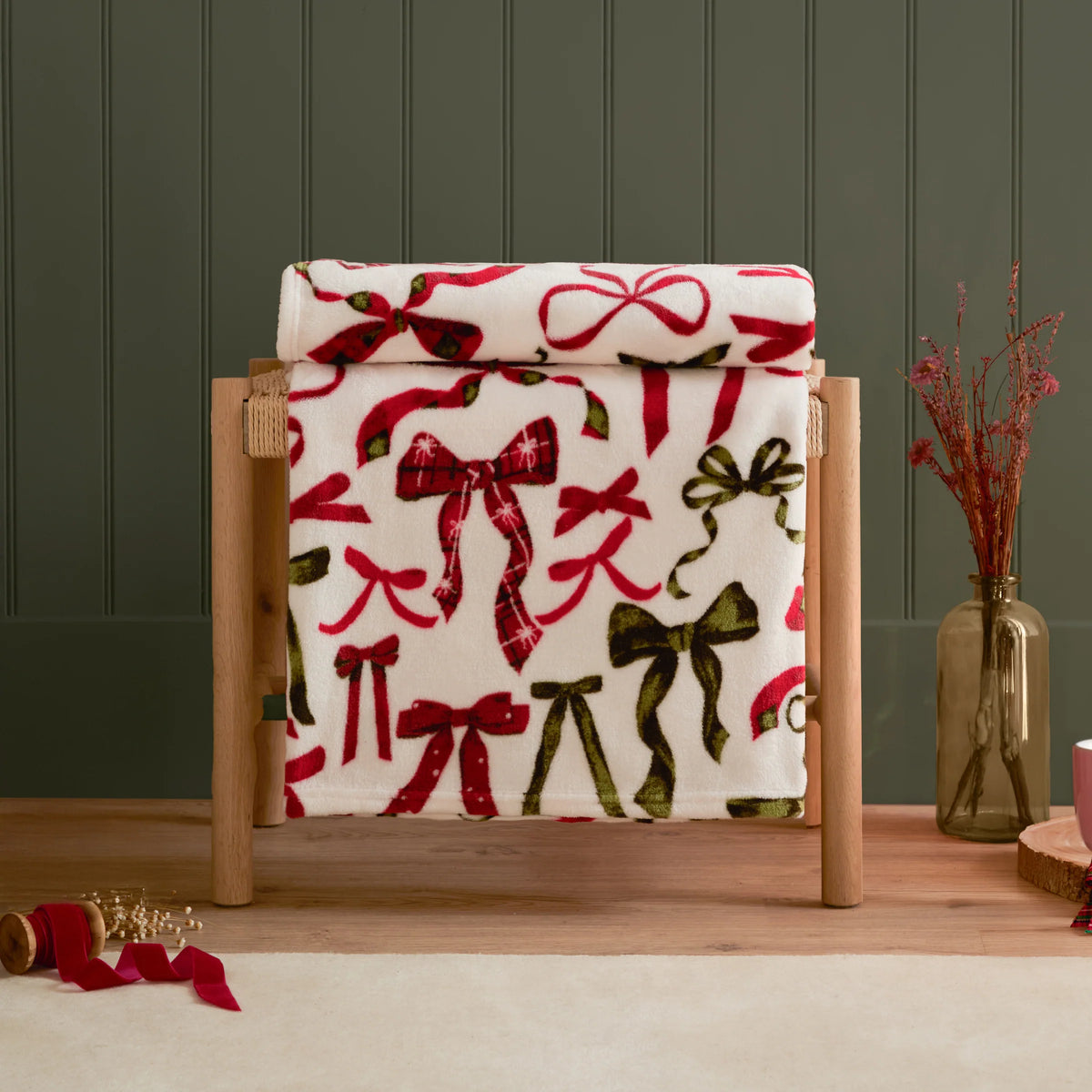 Festive bows print throw rolled and draped over a wooden bench stand against a dark green panelled wall, with a vase of dried pink flowers and a spool of red ribbon on the floor nearby – Kumperi