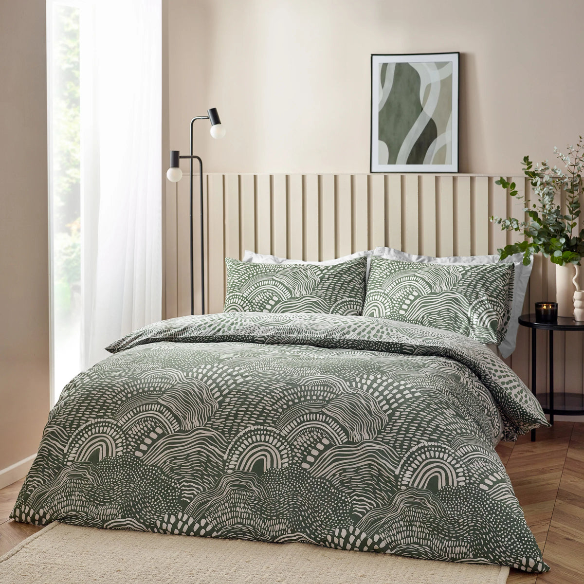 Angled view of the bed dressed in the olive abstract wave duvet cover set with matching pillowcases, styled against a cream slatted headboard with a floor lamp, a framed wave artwork and a eucalyptus arrangement on a side table – Kumperi