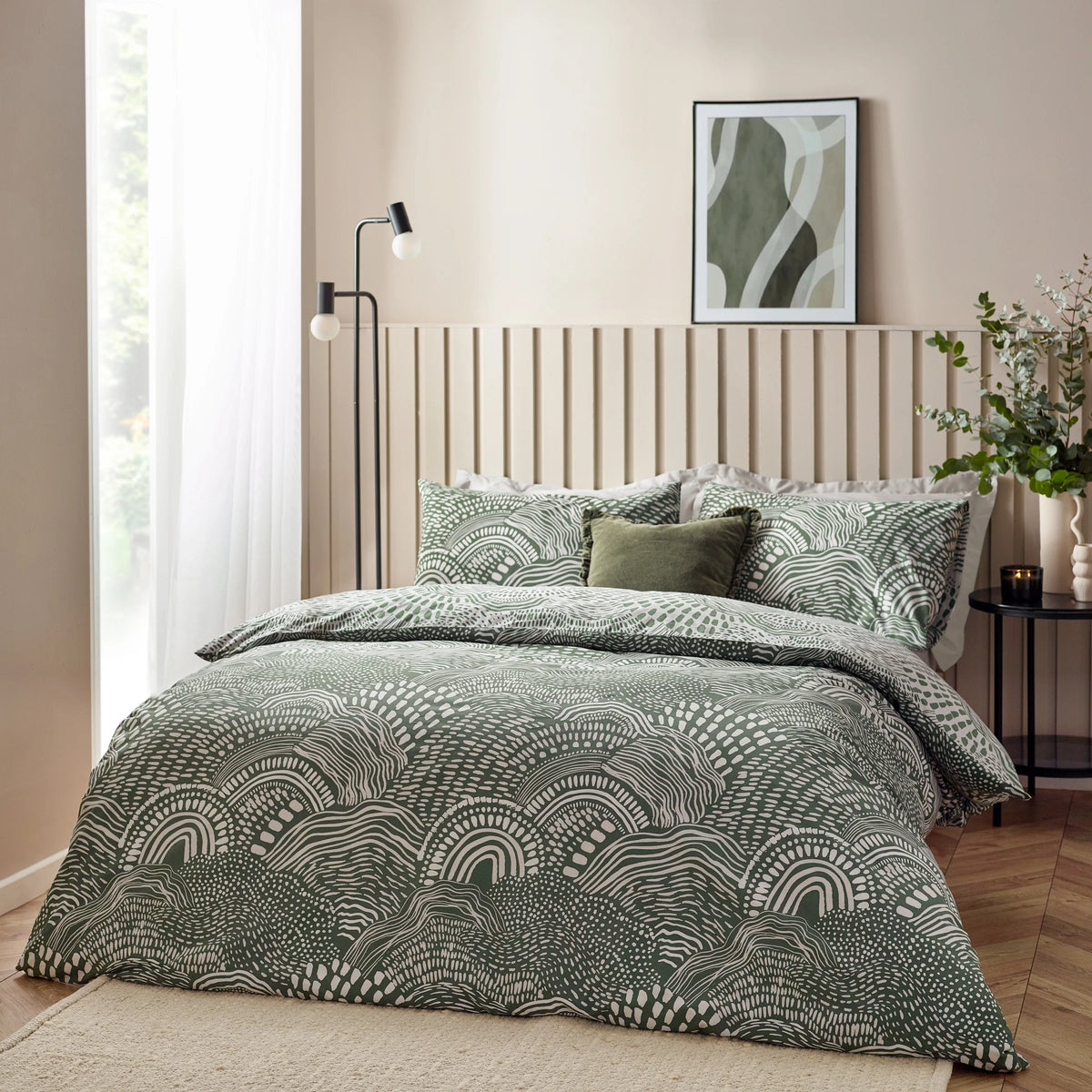 Bed dressed in the olive abstract wave print duvet cover set with matching pillowcases and a plain olive velvet cushion, styled against a cream slatted headboard with a floor lamp, a framed abstract wave artwork and a vase of eucalyptus on a round black side table with a lit candle – Kumperi