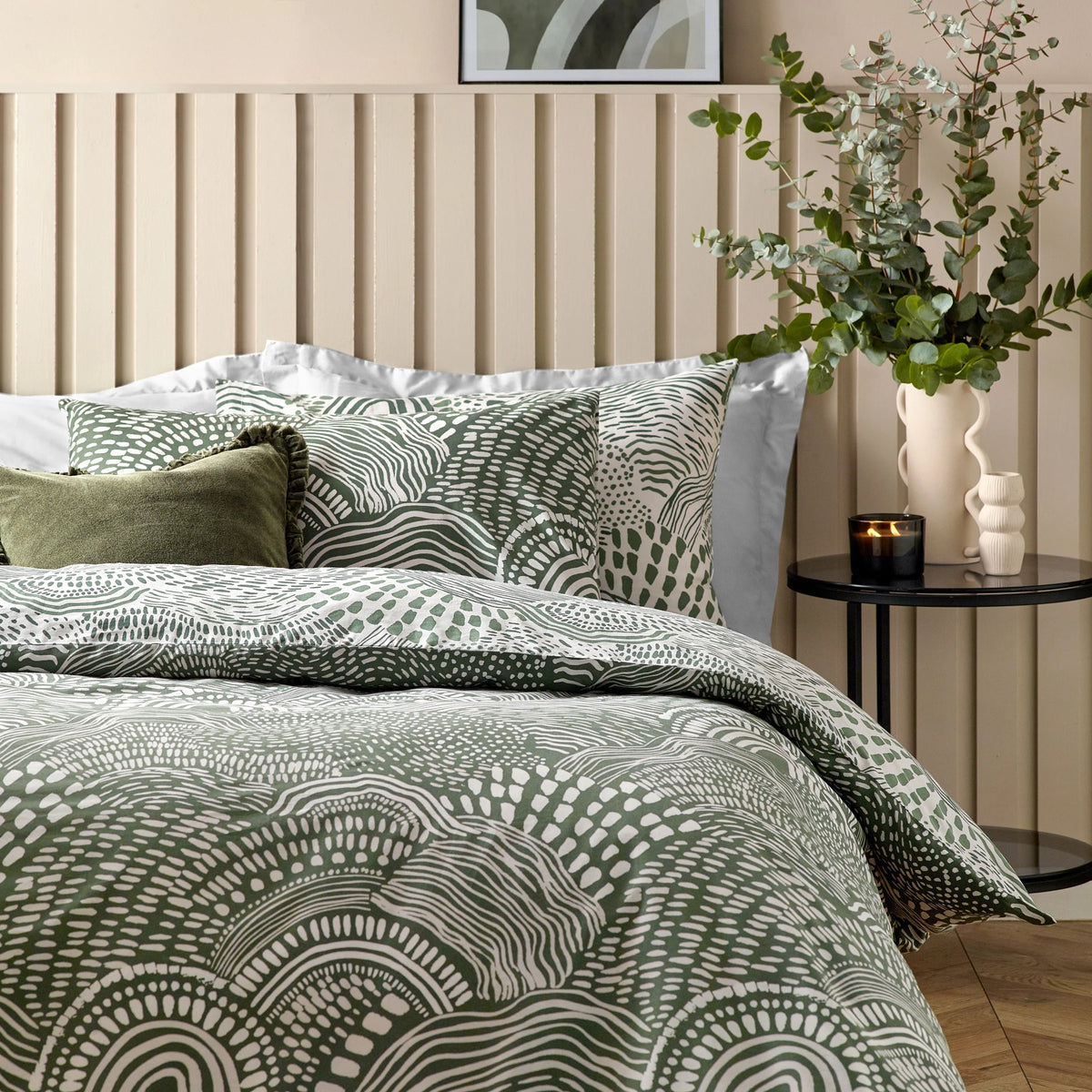 Close view of the bed against a cream slatted headboard showing the olive abstract print pillowcases layered with a plain olive velvet cushion and a ruffled dark green cushion, with a framed wave artwork above and a eucalyptus arrangement on a side table – Kumperi