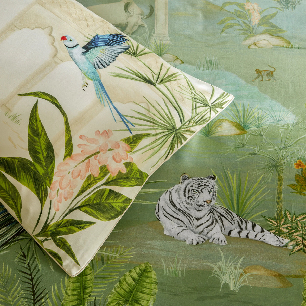 Close-up of a cream pillowcase corner with a jungle print showing a blue and green flying parakeet, palm fronds, pink tropical blooms and a stone archway, set against a wider matching green jungle scene featuring a resting white tiger and a monkey – Kumperi
