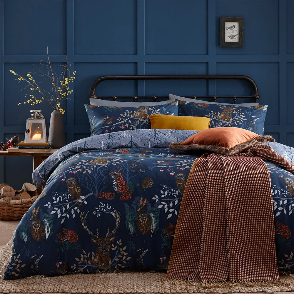 Bed dressed in the navy woodland animal print duvet cover set with matching pillowcases showing foxes, owls, hares and stags among branches, styled with a mustard velvet cushion, a rust waffle throw and a rust faux fur trimmed cushion, set against a dark blue panelled wall with a framed bird print – Kumperi
