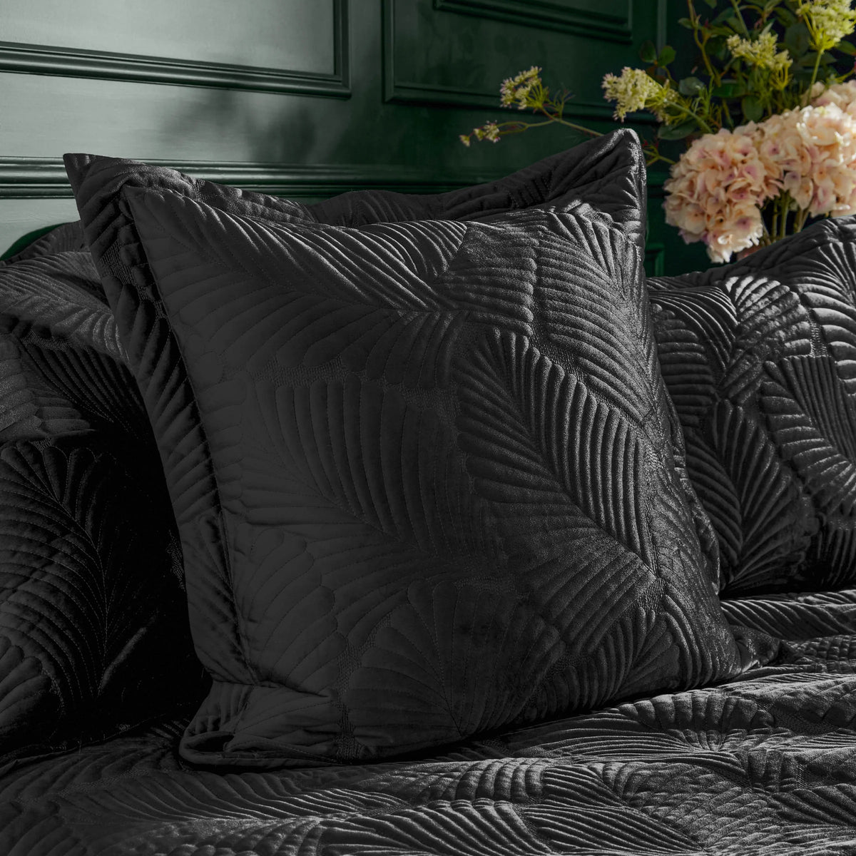Black quilted velvet cushion with an embroidered radiating palm leaf pattern, propped against matching quilted black bedding on a bed set against a dark green panelled wall, styled with a vase of pink hydrangeas and eucalyptus – Kumperi