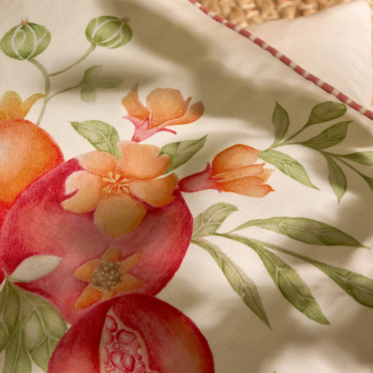Close-up detail of the print showing orange blossom flowers, a split pomegranate revealing its red seeds and trailing olive green leaves against the cream fabric – Kumperi