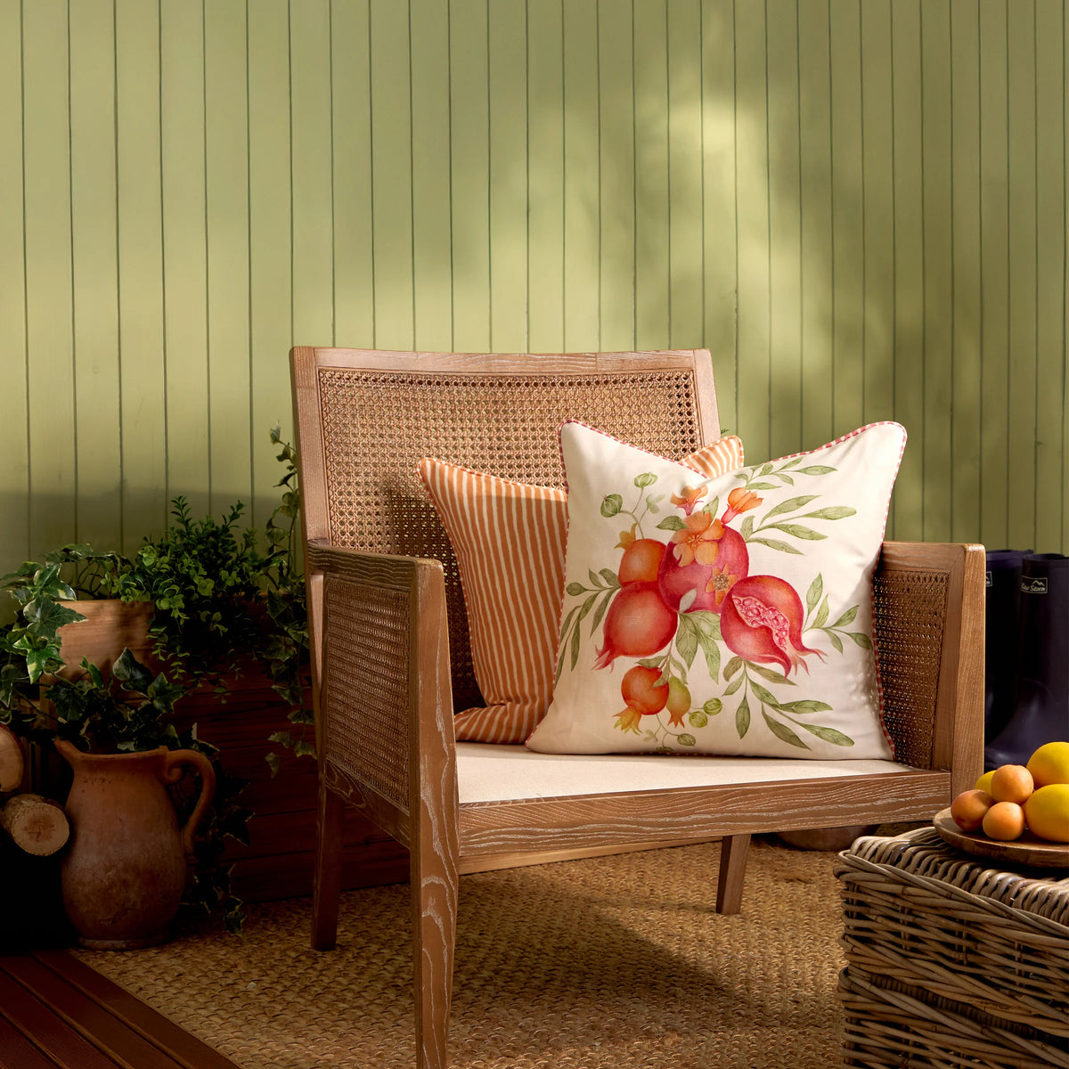 Pomegranate print cushion styled on a rattan bench beside an orange striped cushion, with potted ivy, a terracotta jug, logs and a bowl of citrus fruit nearby against a green wood-panelled wall – Kumperi