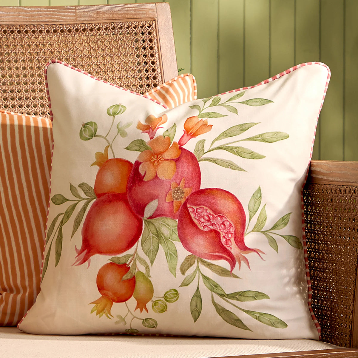 Pomegranate print cushion styled on a rattan-backed wooden chair beside an orange and white striped cushion, the checked piped edge visible along the seam – Kumperi