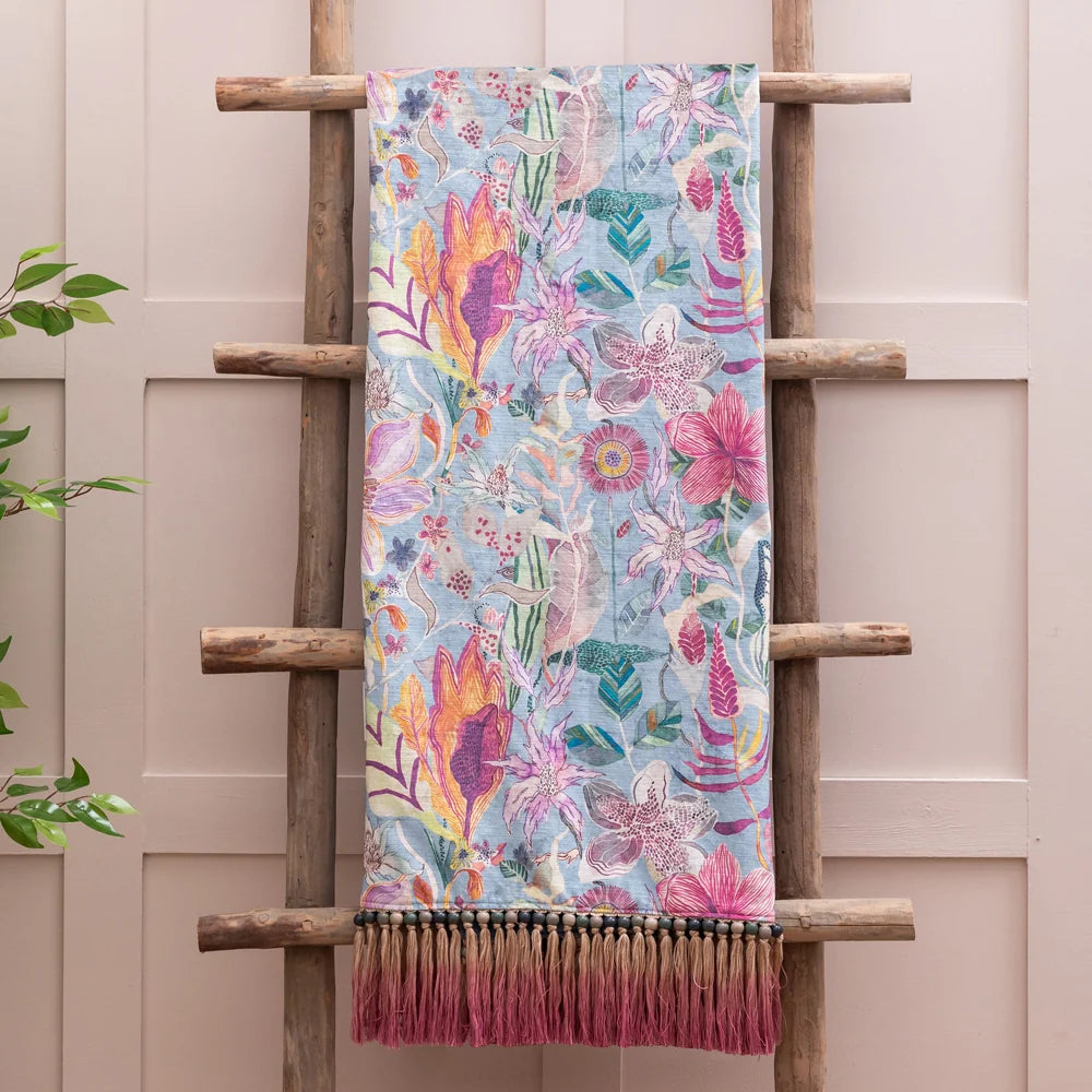 Floral printed throw hanging over a wooden ladder against a pale pink panelled wall, showing a dense watercolour-style pattern of magenta, orange and pink flowers with green and purple foliage on a powder blue ground, finished with a tan and pink beaded fringed trim – Kumperi