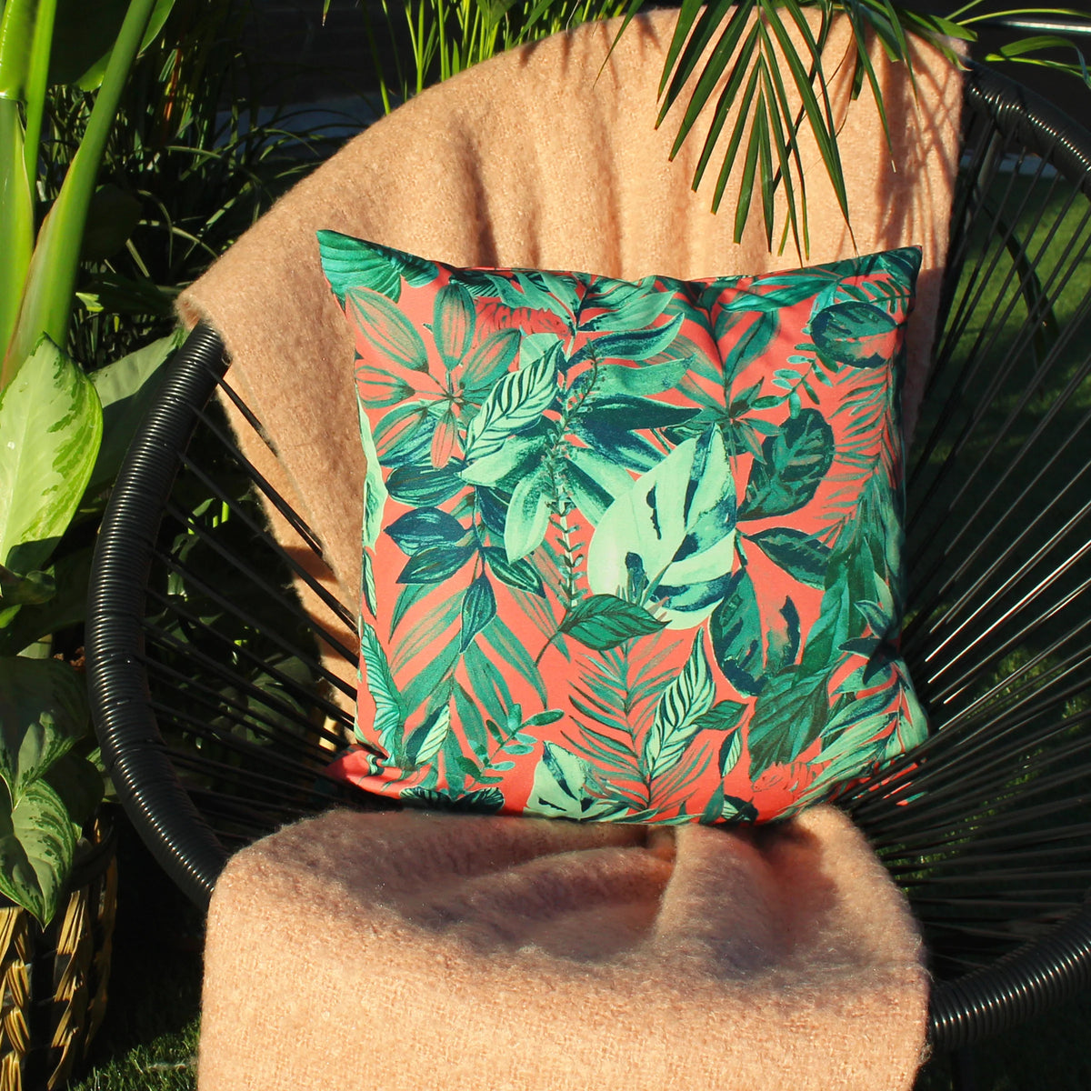 Coral jungle print cushion styled on a black woven outdoor chair draped with a camel-coloured throw, surrounded by real green tropical plants in a sunlit garden – Kumperi