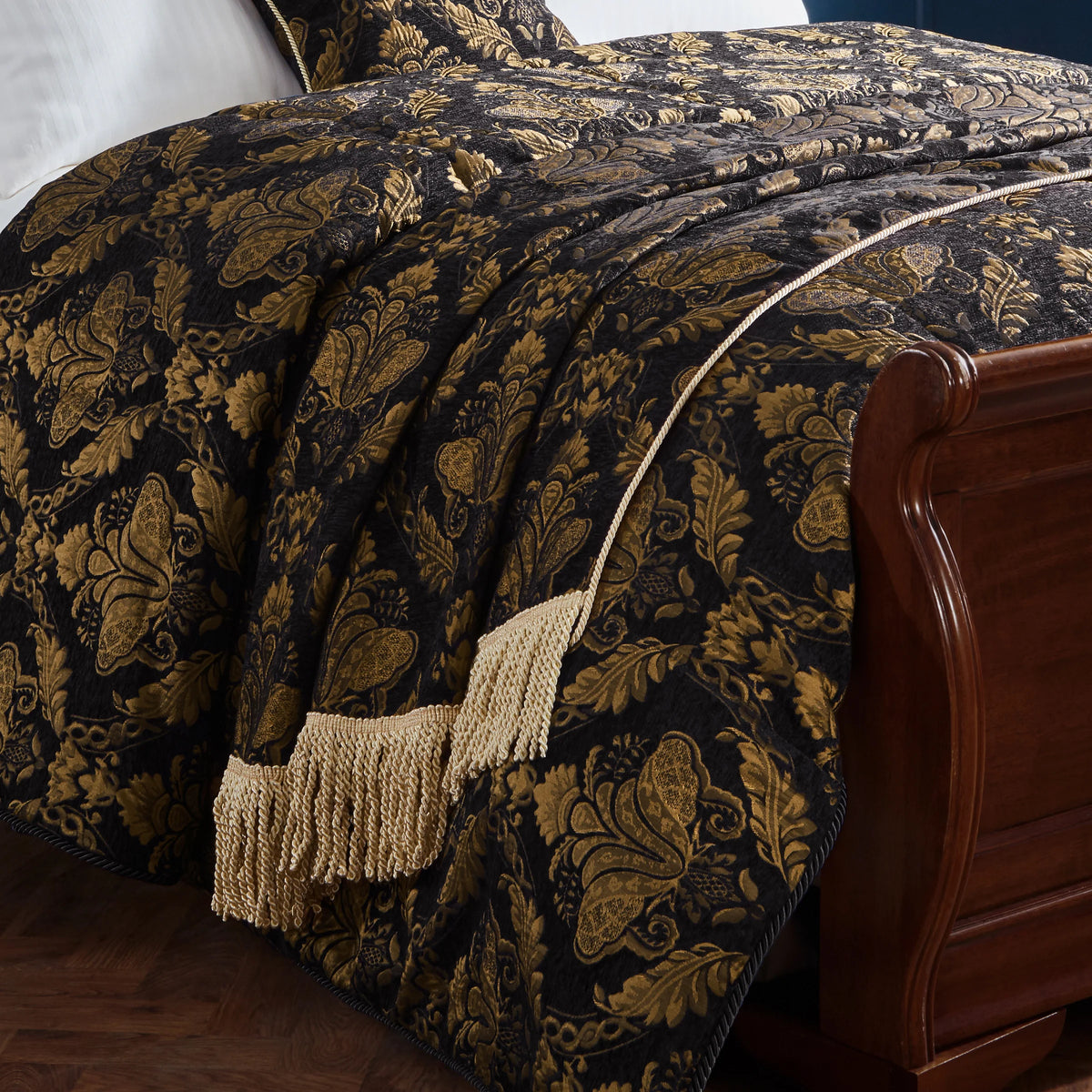 Black and gold jacquard throw draped over a bed with matching cushions, showing the damask pattern and cream fringed trim styled against a dark wooden sleigh bed frame – Kumperi
