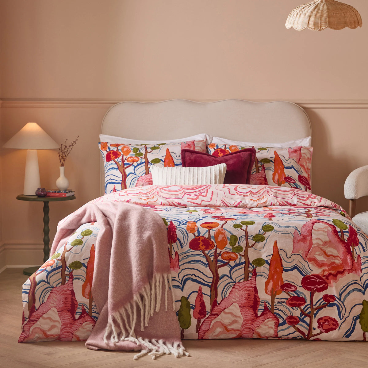Bed dressed in the multicolour painted mountain print duvet cover set with matching pillowcases, layered with a burgundy velvet cushion, a cream ribbed cushion and a pink throw pillow, a pink mohair throw draped over the front, styled against a beige scalloped headboard with a rattan pendant light – Kumperi