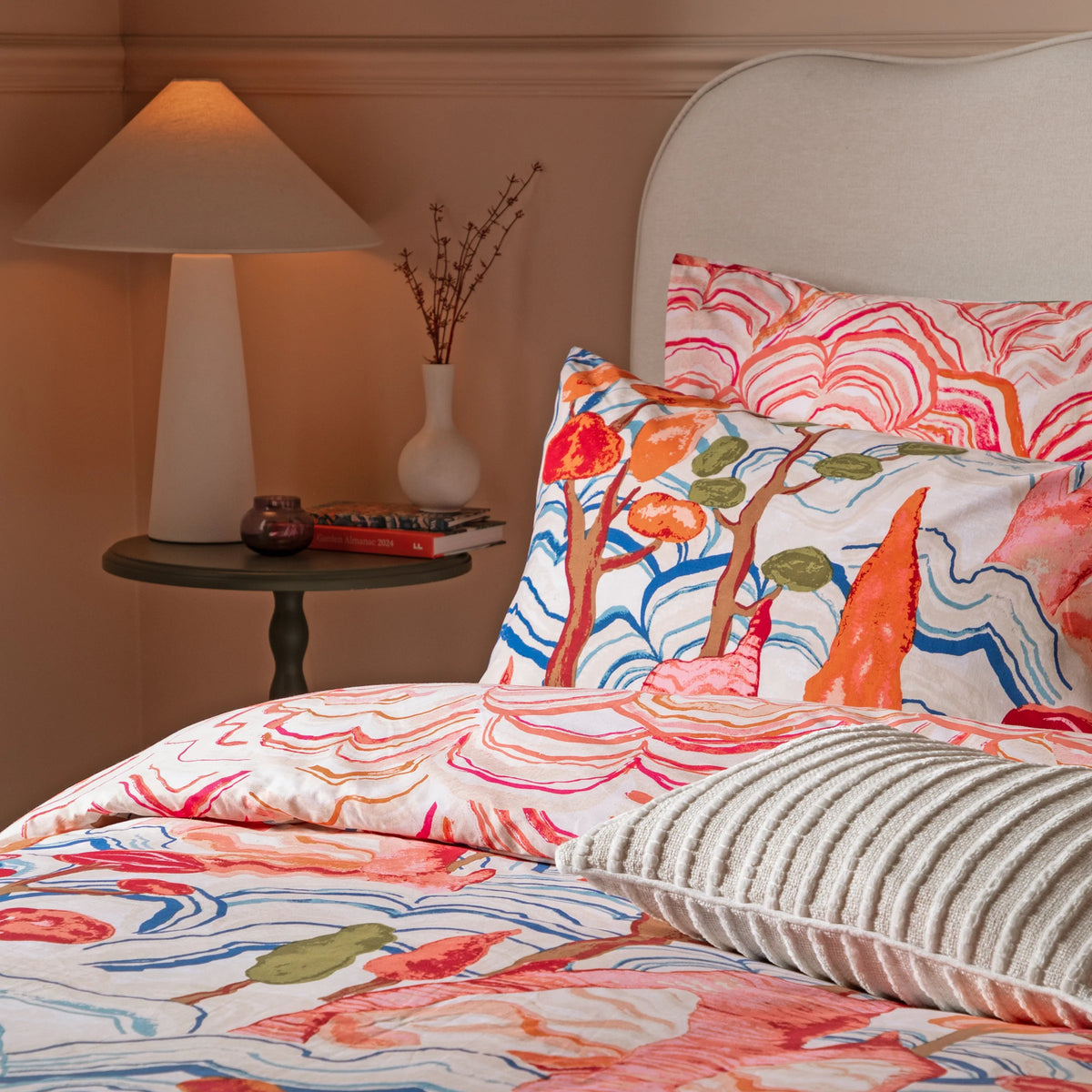 Close view of stacked pillowcases showing both the coral mountain print and the reversible blue and orange tree print, with a cream ribbed cushion in the foreground and a bedside lamp and vase in the background – Kumperi