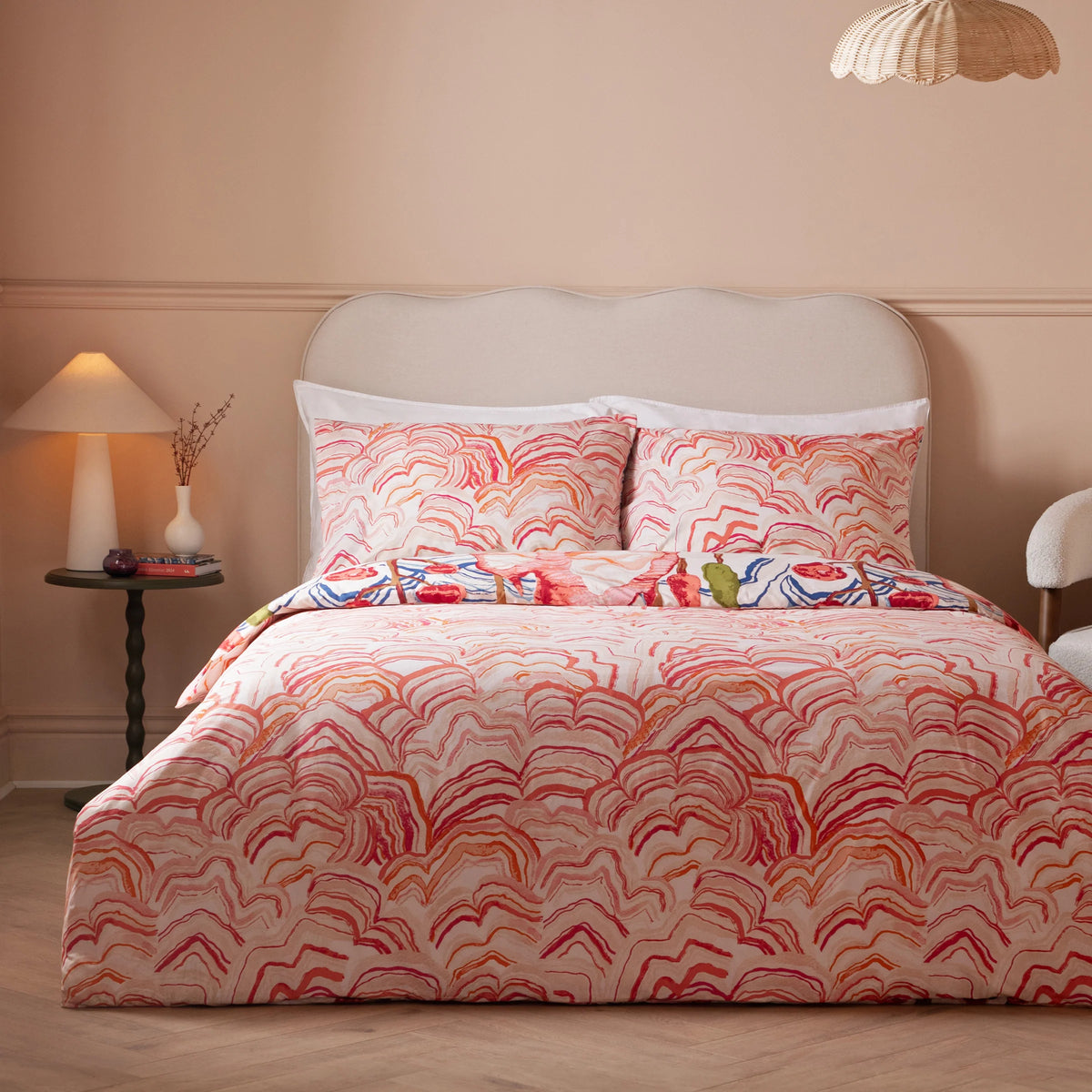 Close-up of the coral and pink mountain print pillowcases stacked together, showing layered wavy ridge lines in fine detail against a beige scalloped headboard – Kumperi
