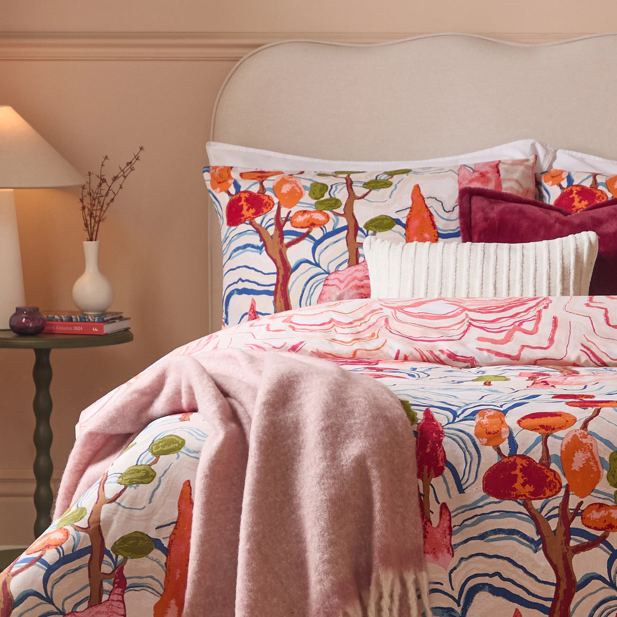 Close-up of the reversible tree and mountain print pillowcase in blue, orange and olive tones, layered with a burgundy velvet cushion and a cream ribbed cushion, with the coral mountain print duvet cover visible below – Kumperi