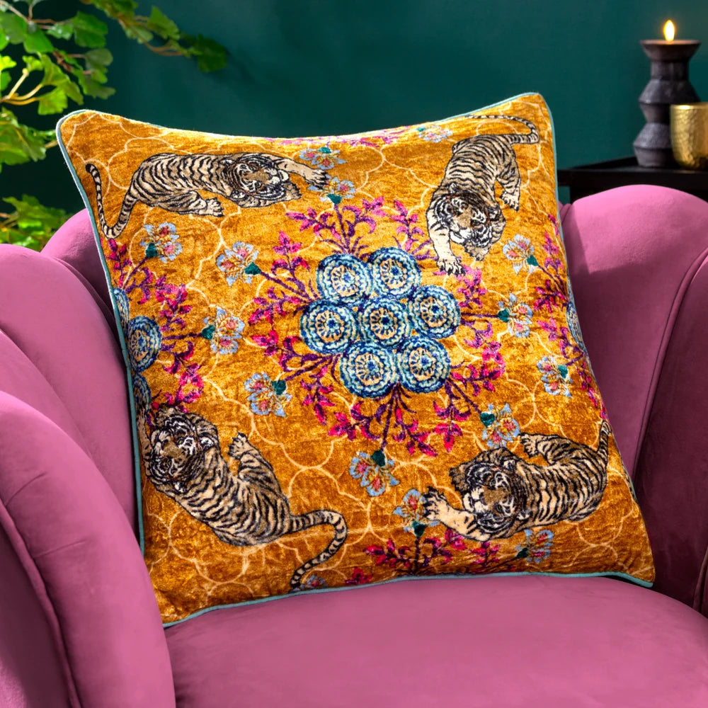 Gold tiger print velvet cushion with teal piping styled on a pink velvet armchair, set against a dark teal wall with a lit candle and green foliage nearby – Kumperi