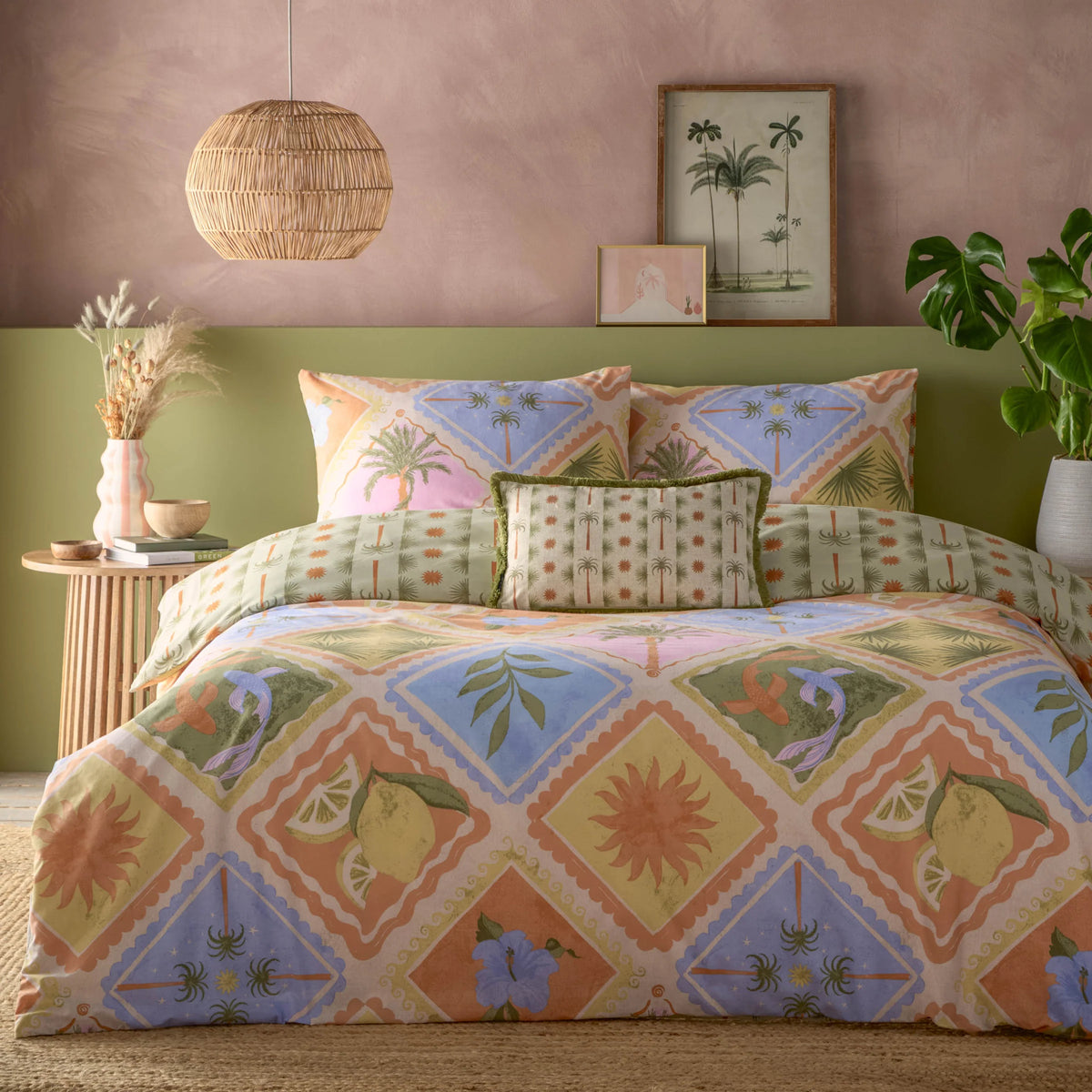 Bed dressed in the multi tropical patchwork print duvet cover set with matching pillowcases showing palm trees in diamond panels, a sage green reversible panel, and a fringed palm tree print bolster cushion, styled against a dusty pink wall and olive green headboard with framed palm tree artwork, a rattan pendant light and a monstera plant – Kumperi