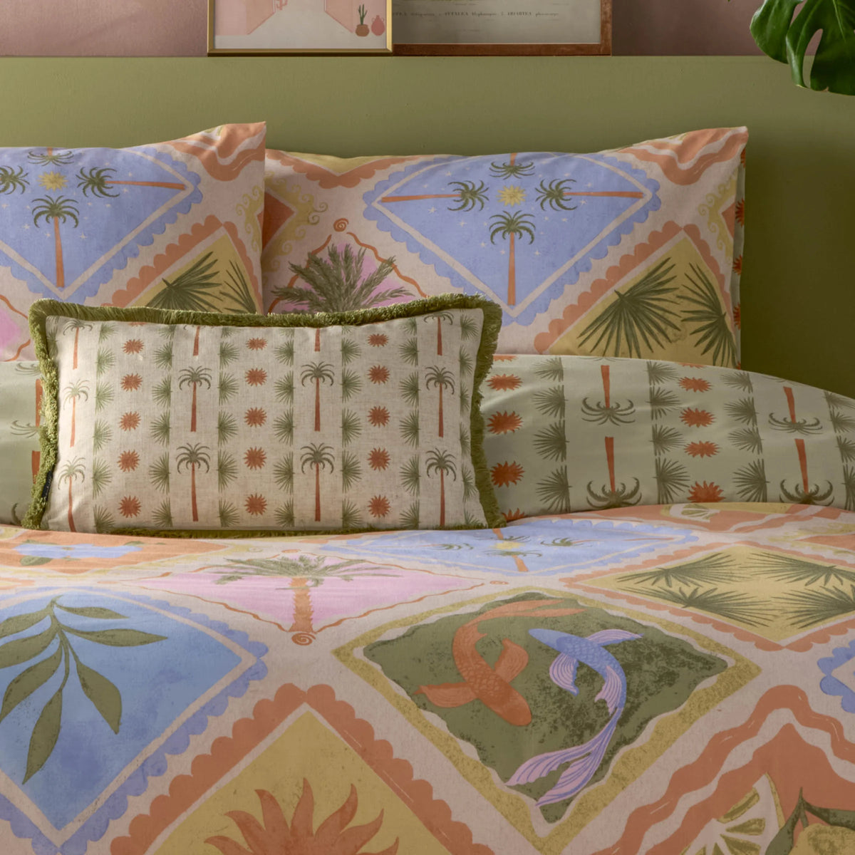 Close view of stacked pillowcases in the tropical patchwork print and sage green palm tree reverse, with a fringed palm tree bolster cushion in front, showing koi fish and palm tree motifs on the duvet cover below, set against an olive green headboard – Kumperi