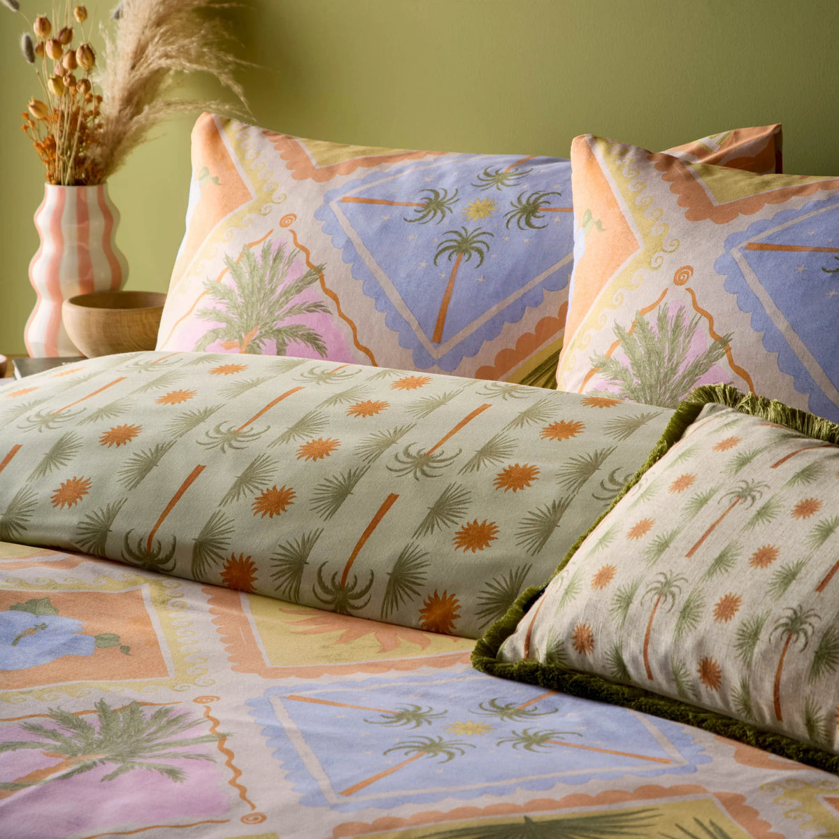 Close-up of two tropical patchwork print pillowcases showing palm trees and sunburst motifs in blue and coral diamond panels, layered over the sage green reversible fabric and a fringed palm tree print bolster cushion, styled beside a striped vase with dried flowers – Kumperi